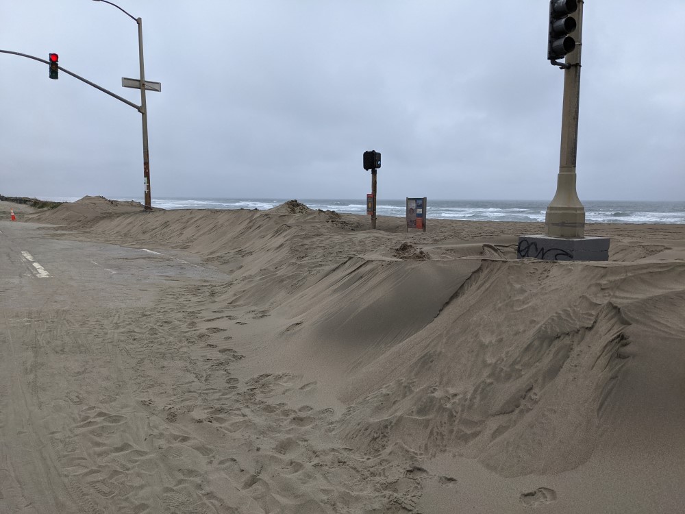 Sand dunes that have shifted to swallow the road and road signs.