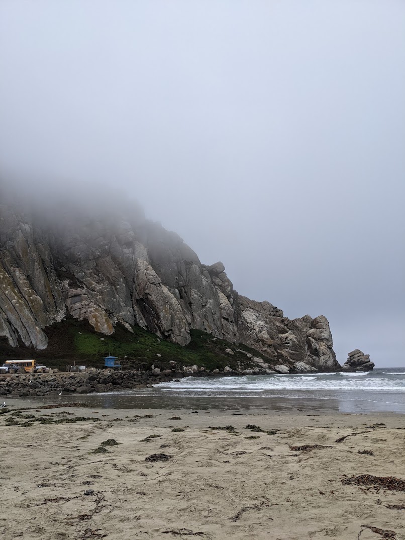 A rocky hill by the sea, a beach in the foreground, a small blue lifeguard station in the distance and a school bus. The top of the hill is shrouded in fog.