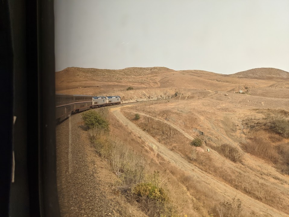 Out the window of the train, seeing the train curve to the right, surrounded by brown dry hills.