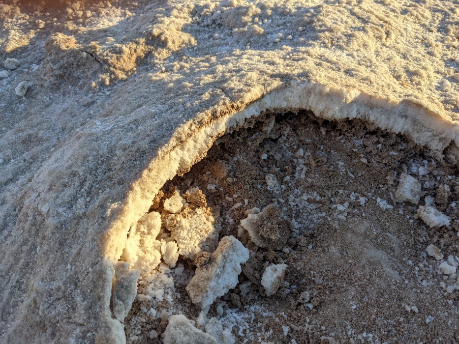 Close up of the ground. Packed, it looks like snow, but a hole has been broken through showing a rust of salt crystals an inch thick, with dirt beneath.